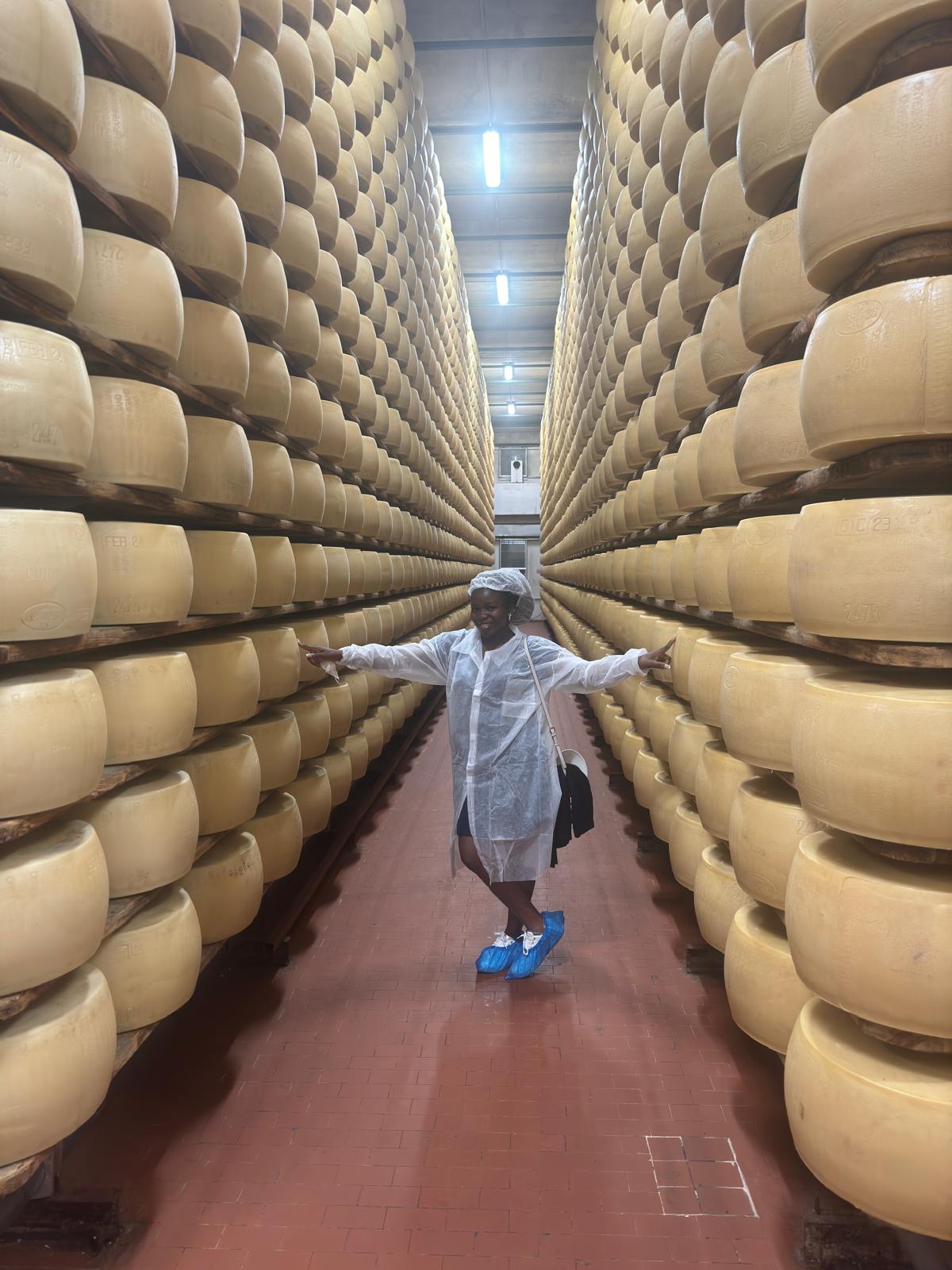 Temi standing arms outstretched in a Parmigiano-Reggiano ageing cave surrounded by stacked cheese wheels